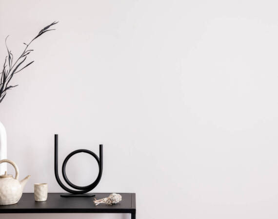 Aesthetic interior of living room with copy space, white wall, black table, candlestick, vase with dried flowers, pitcher, and elegant personal accessories. Stylish home decor. Template.